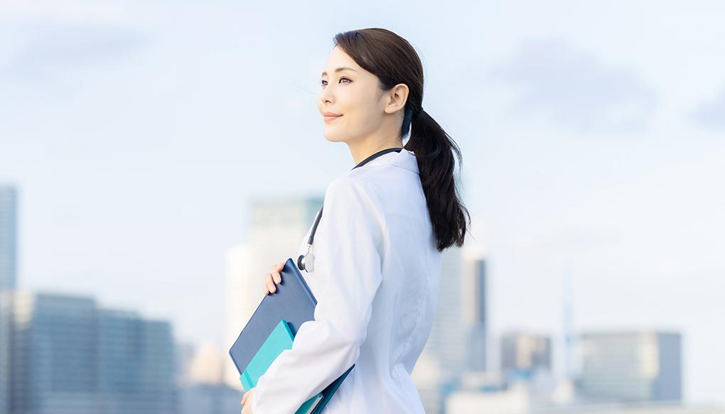 都市部の青空を背に立つ白衣の女性医師
